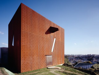 Image of a rust colored, metal clad structure on a hilly site in Shiogama, Japan, with a view of the Pacific Ocean.