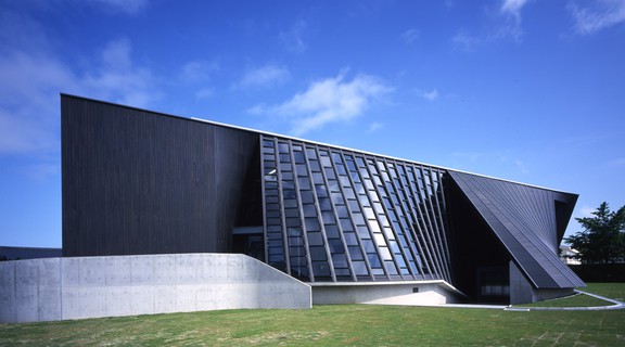 Exterior view of Reihoku Community Hall, a grey slanted building with windows along the middle of the facade, surrounded by green lawn.