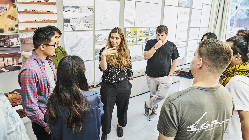 Female faculty member speaking to a group of students against a backdrop of student work pinned up on the wall