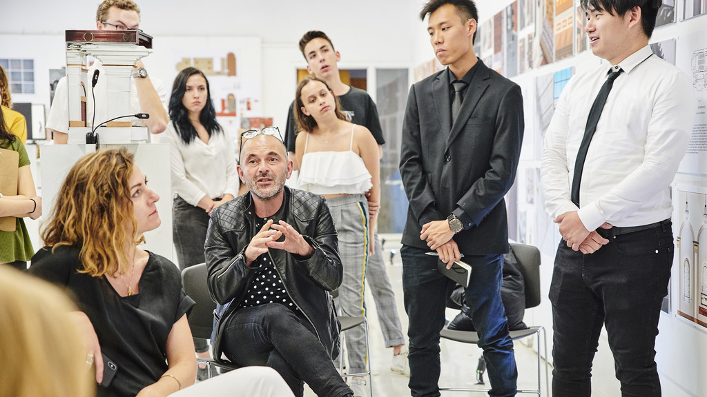 Male faculty member speaking to a group of students against a backdrop of student work pinned up on the wall