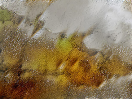 Aerial photograph of dried lake bed with rippled surface like a lake in the wind. Underneath the rippled surface can be seen a mixture of green, yellow, and rust pigments.